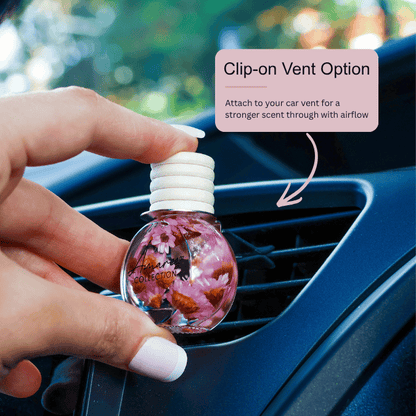 A hand holds a small round car air freshener bottle with pink floral contents and a white clip-on cap, positioned near a car vent. A pink text box reads &