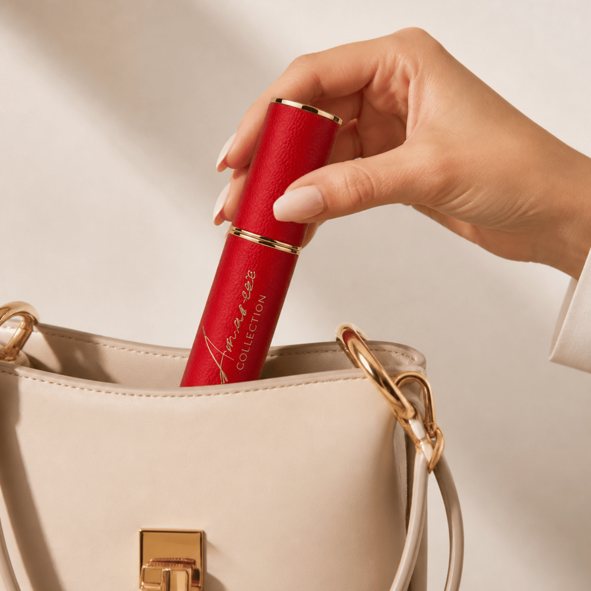 A hand with manicured nails holds a red lipstick tube with gold trim, inserting it into a beige handbag with gold hardware.