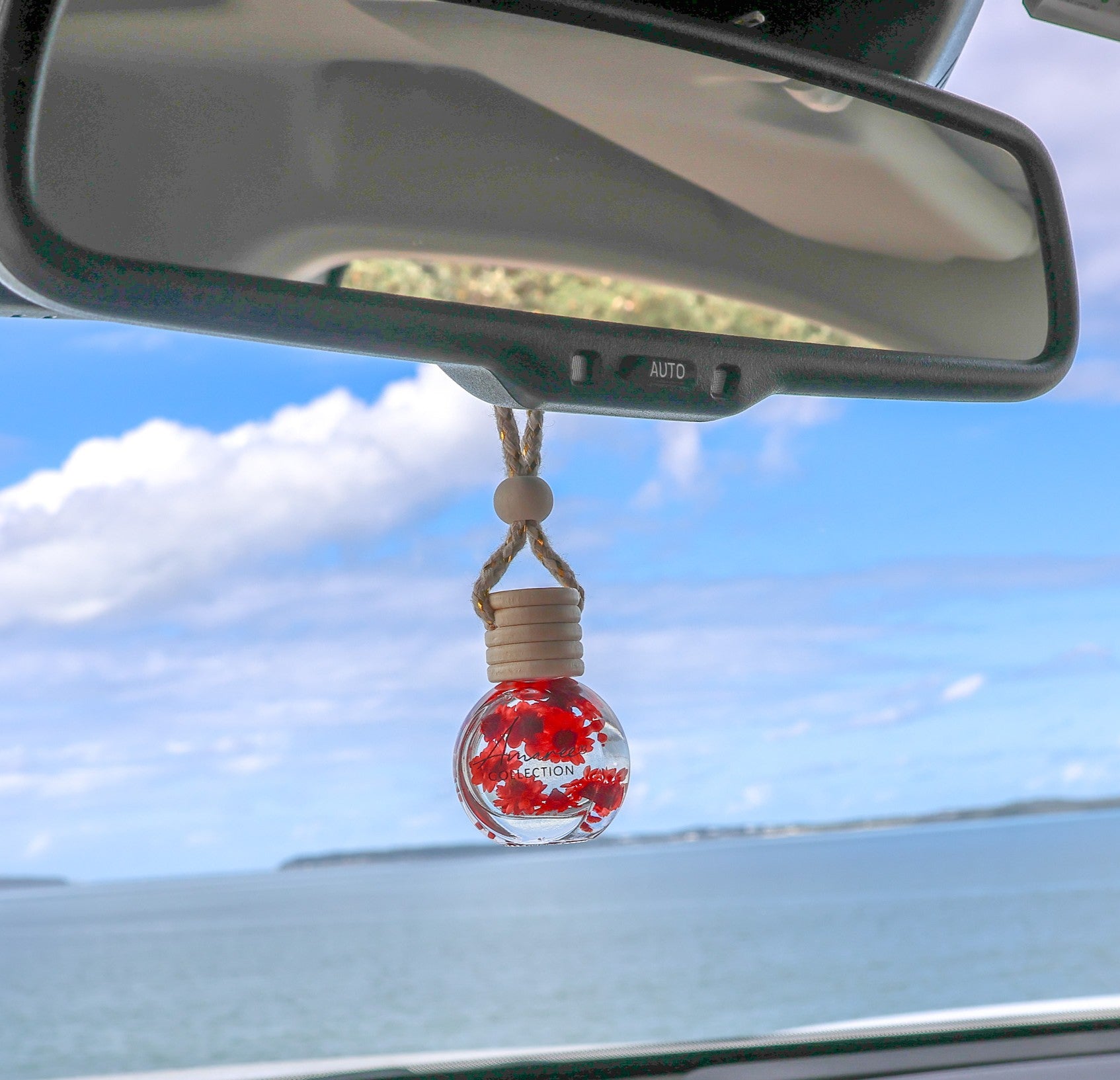 A car air freshener ornament shaped like a glass bottle with red flowers inside, hanging from a rope under a rearview mirror in a car with a blue ocean and sky background.