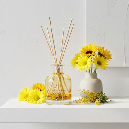 A diffuser with reeds and a small vase of yellow flowers on a white surface.