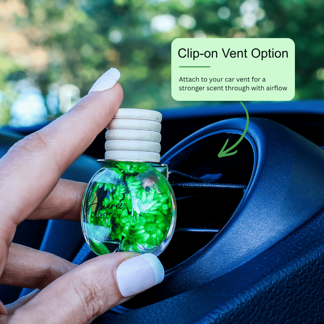A hand with white manicured nails holds a clear glass car air freshener bottle filled with bright green botanical pieces. The bottle has a white ribbed cap and is positioned near a car air vent, with a green text box overlay explaining the clip-on vent option.