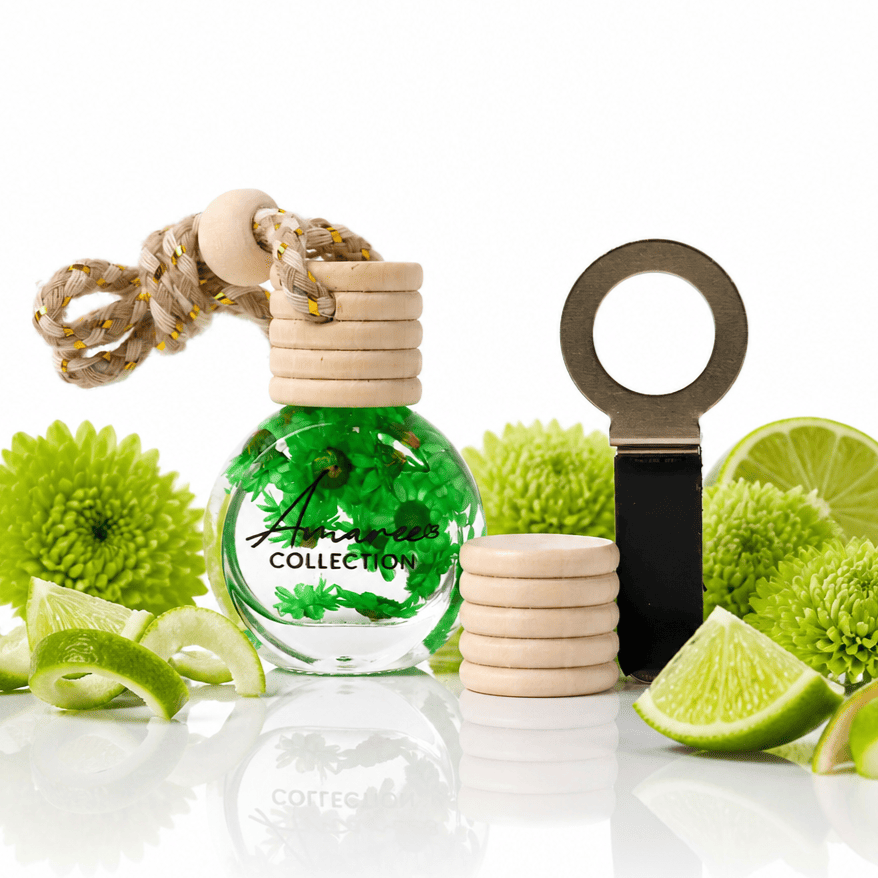 A green glass car air freshener bottle with a wooden diffuser cap and rope loop, styled with lime slices, green flowers, and a metal key ring on a white reflective surface.
