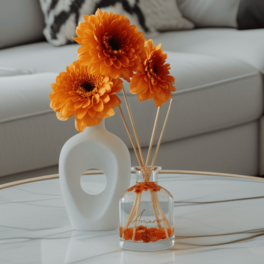 Orange flowers in a white vase and diffuser on a coffee table with a blurred sofa in the background