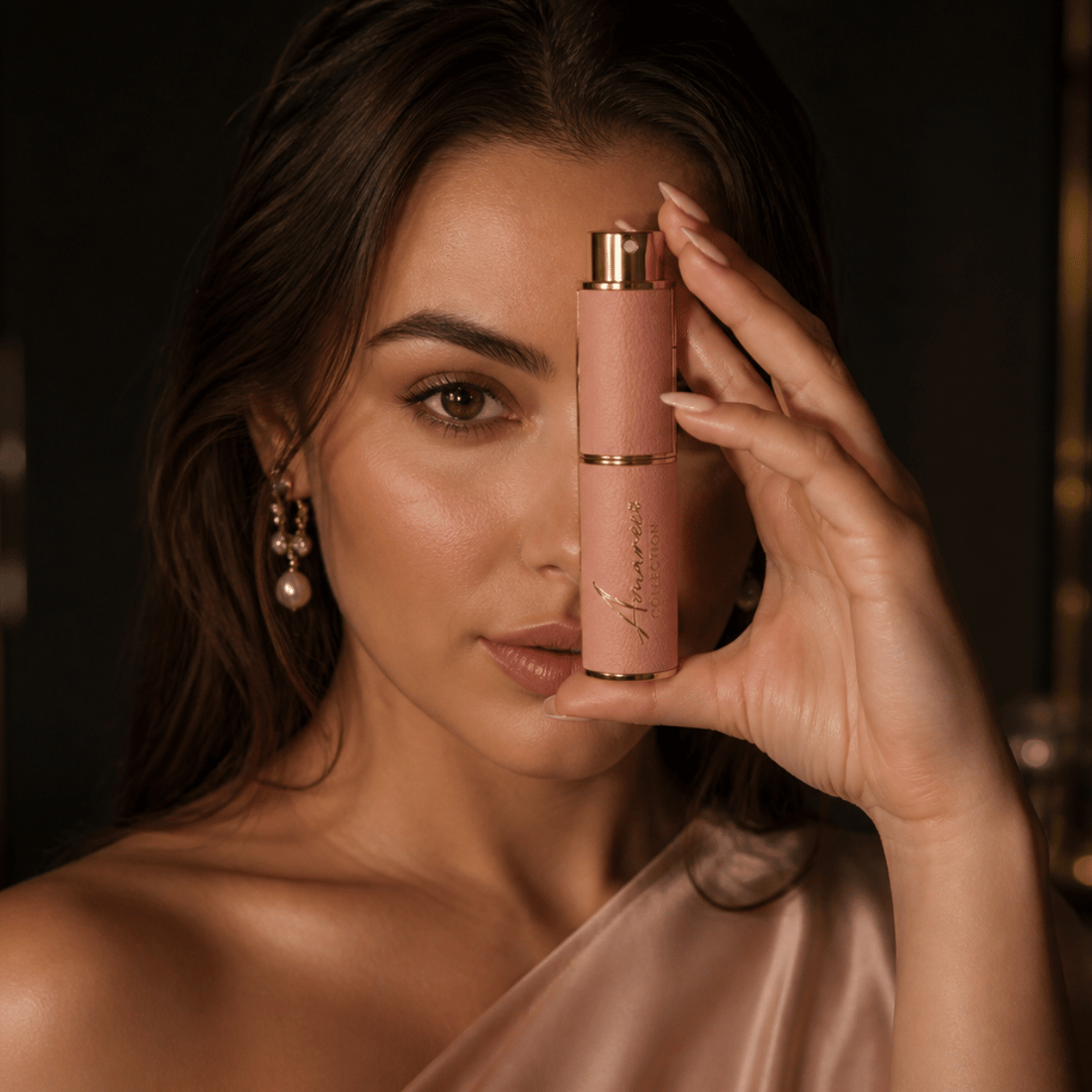 A woman holds a pink cylindrical cosmetic bottle with a gold cap near her face against a dark, softly lit background.