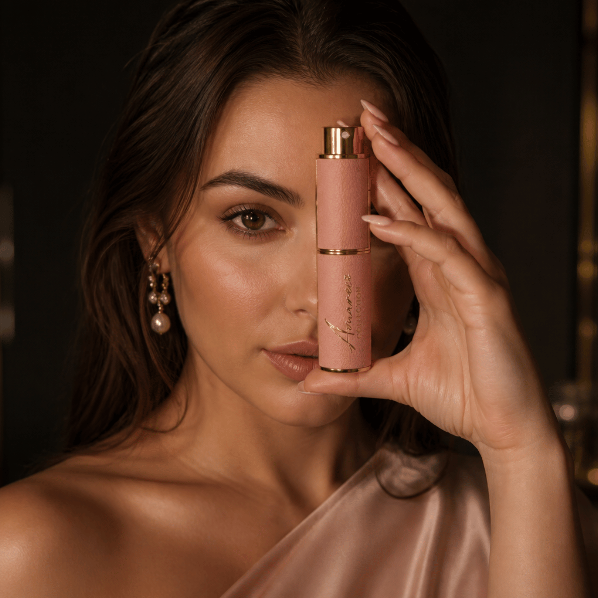 A woman holds a pink cylindrical cosmetic bottle with a gold cap near her face against a dark, softly lit background.