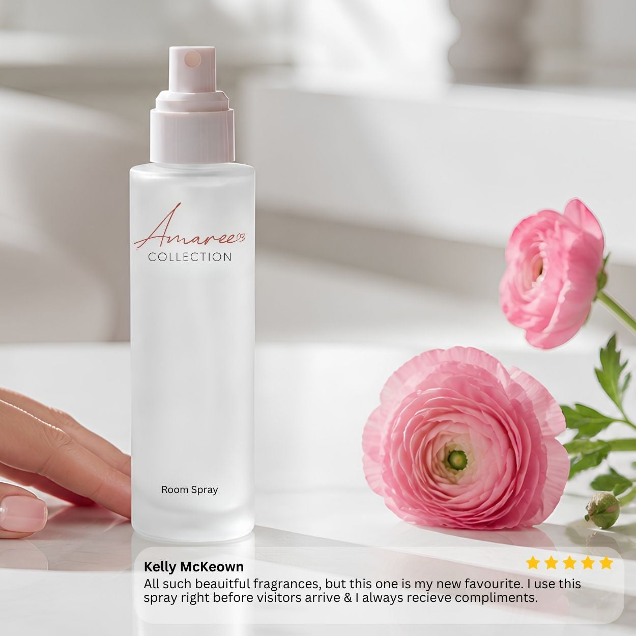 A frosted white room spray bottle from Amaree Collection stands on a white surface next to pink ranunculus flowers. A hand is visible on the left side of the bottle.