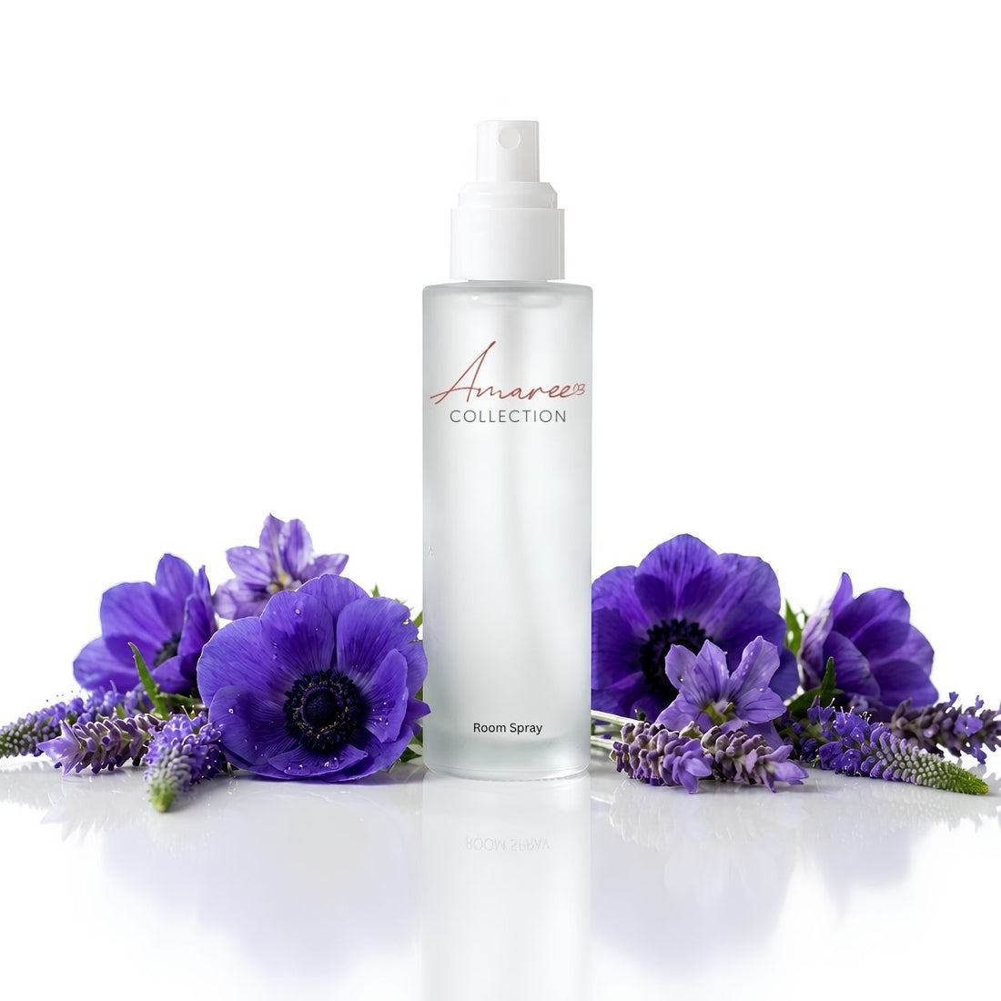 A clear bottle of Amaree Collection Room Spray with a white pump, styled with purple flowers and lavender sprigs on a reflective white surface against a white background.