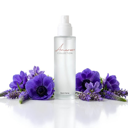 A clear bottle of Amaree Collection Room Spray with a white pump, styled with purple flowers and lavender sprigs on a reflective white surface against a white background.
