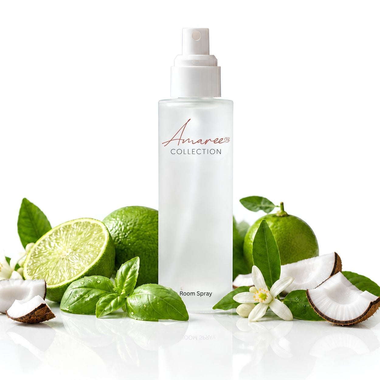A frosted glass room spray bottle from Amaree Collection, surrounded by fresh green limes, basil leaves, white flowers, and coconut halves on a reflective white surface.