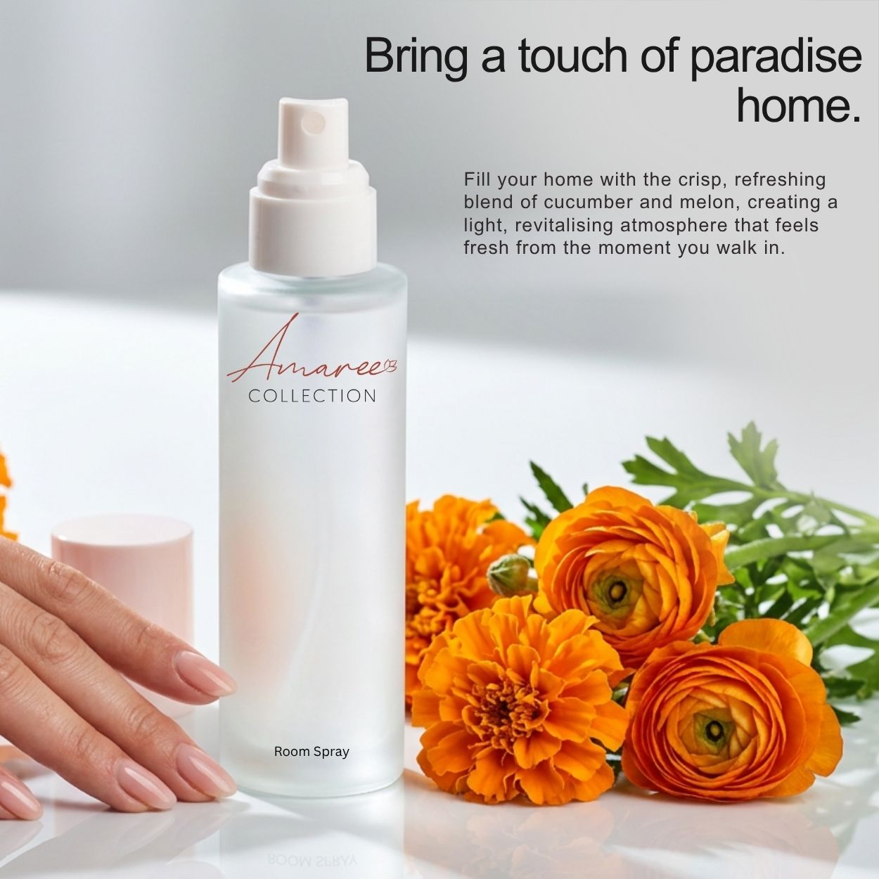 A frosted glass room spray bottle from Amaree Collection stands on a white surface beside a hand with pale pink manicured nails and bright orange flowers. Text at the top reads, &