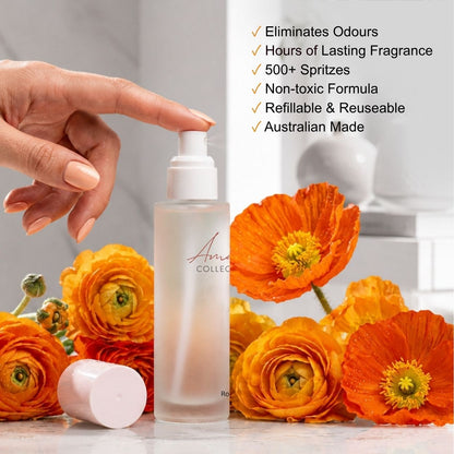 A frosted glass air freshener spray bottle with a white pump, shown with a hand pressing the nozzle. Bright orange flowers surround the bottle, and a white cap lies on its side.
