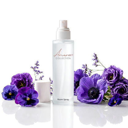A frosted glass room spray bottle from Amaree Collection stands upright on a reflective white surface, surrounded by purple flowers and a small white candle in a white pot.
