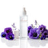 A frosted glass room spray bottle from Amaree Collection stands upright on a reflective white surface, surrounded by purple flowers and a small white candle in a white pot.