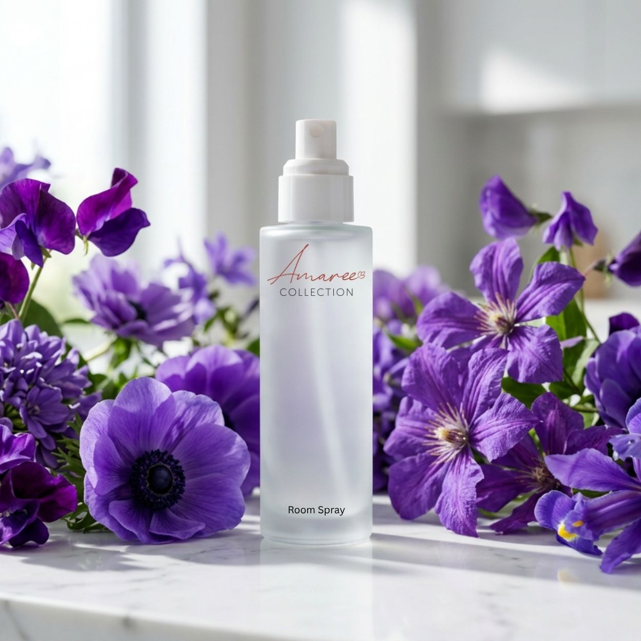A frosted glass room spray bottle from Amaree Collection stands on a white marble surface, surrounded by vibrant purple flowers against a soft, bright background.