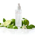 A frosted glass room spray bottle from Amaree Collection, surrounded by fresh green limes, basil leaves, and white jasmine flowers on a reflective white surface.