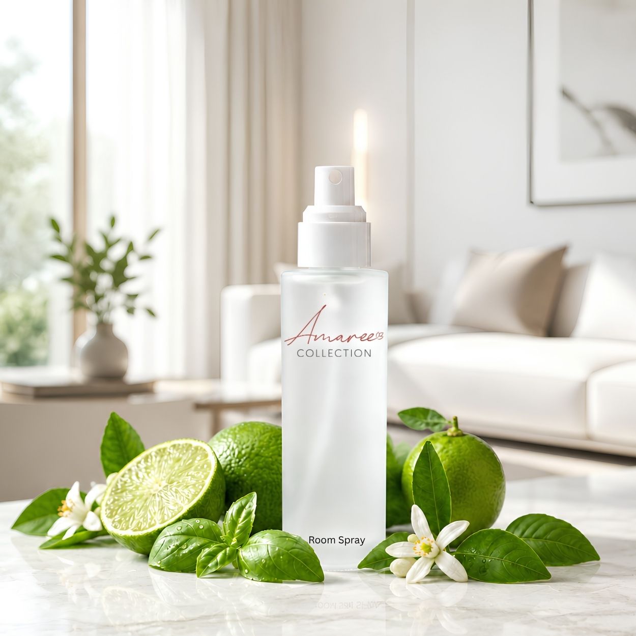 A white room spray bottle from Amaree Collection stands on a marble surface surrounded by fresh limes, basil leaves, and white flowers, with a softly blurred living room background.