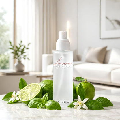 A white room spray bottle from Amaree Collection stands on a marble surface surrounded by fresh limes, basil leaves, and white flowers, with a softly blurred living room background.