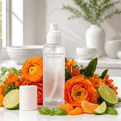 A frosted glass room spray bottle from Amaree Collection stands on a white surface surrounded by bright orange flowers, orange slices, lime wedges, and green leaves, with a small white cylindrical container beside it.