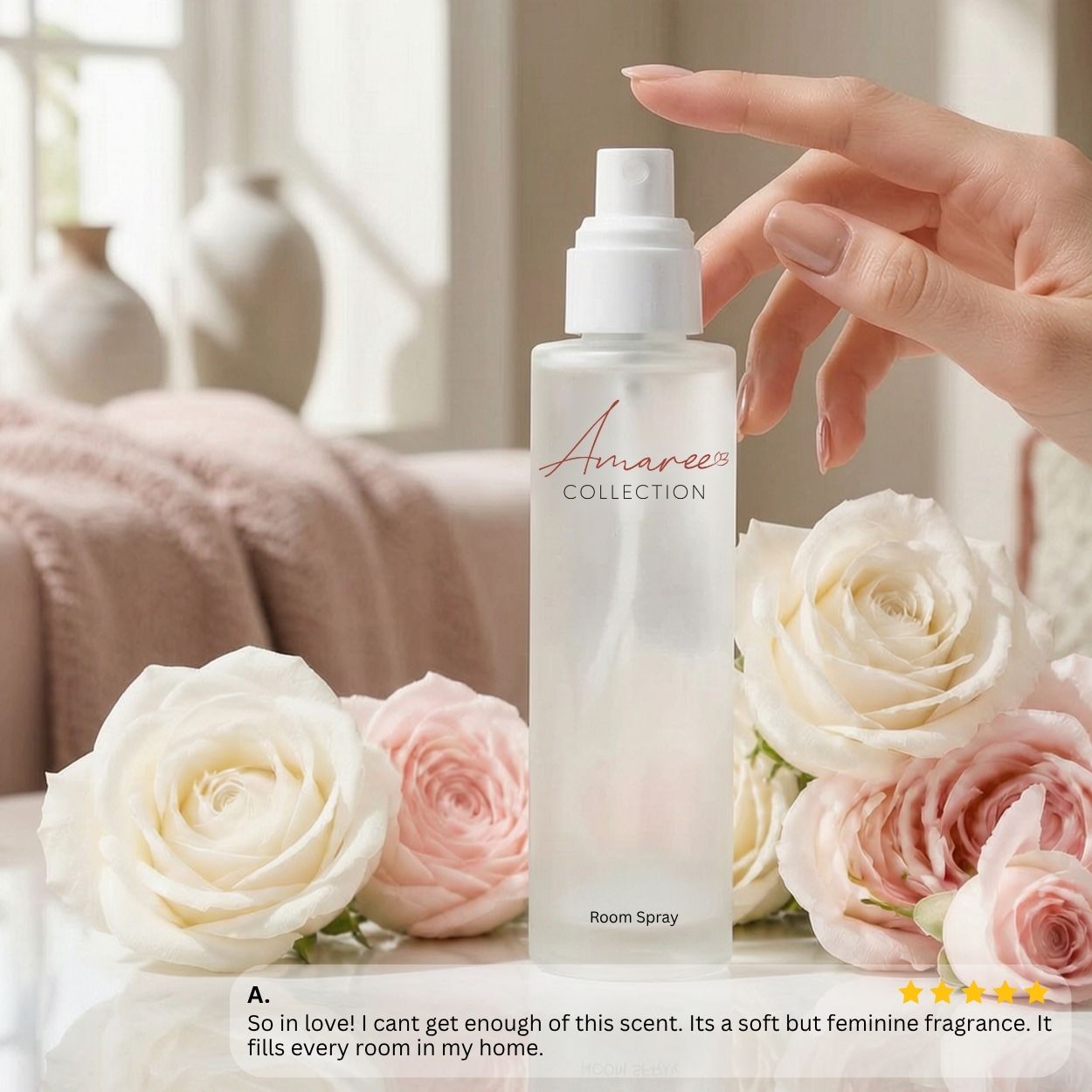 A frosted glass room spray bottle from the Amaree Collection stands on a tabletop surrounded by white and pink roses. A hand with manicured nails is reaching toward the bottle.