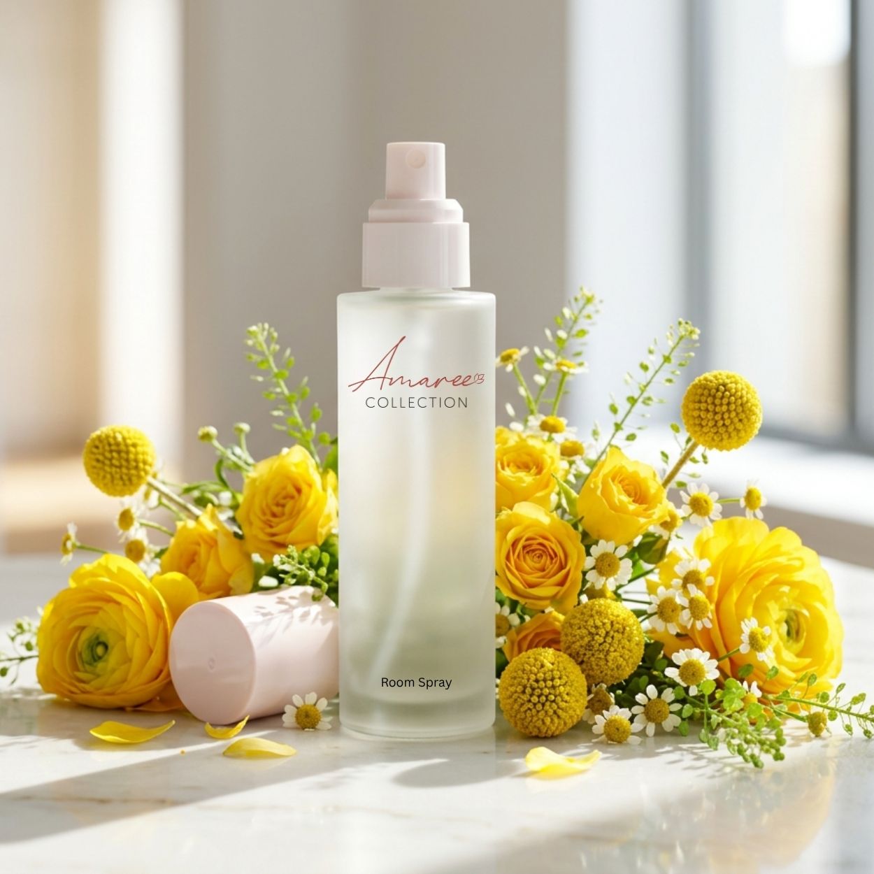A frosted glass room spray bottle with a pale pink pump stands on a light surface, surrounded by yellow flowers and green stems. A matching pale pink cap lies beside the bottle.