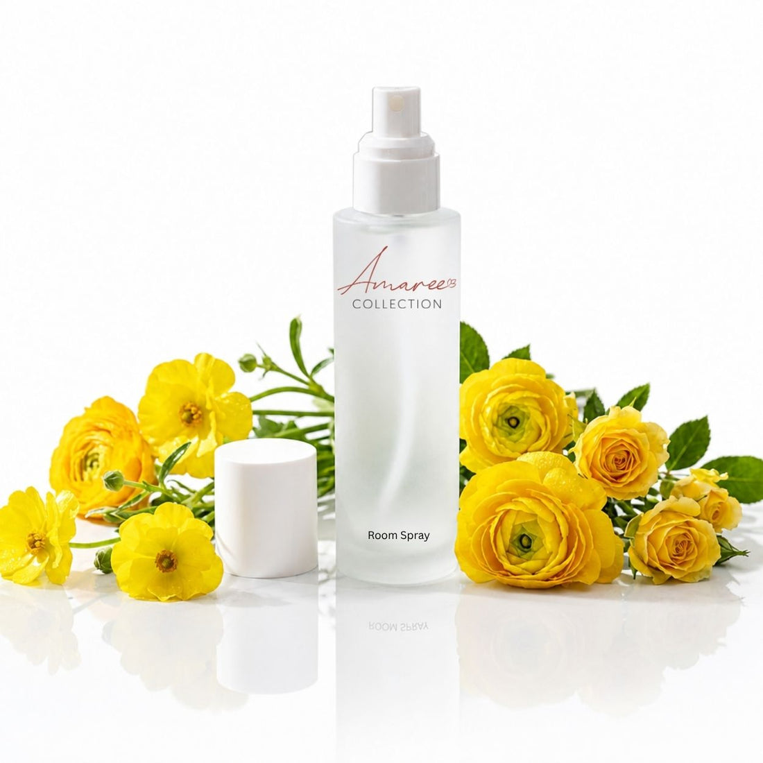 A white bottle of Amaree Collection Room Spray stands on a reflective white surface, flanked by bright yellow flowers and green leaves. The bottle has a white spray top and the brand name &