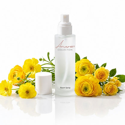 A white bottle of Amaree Collection Room Spray stands on a reflective white surface, flanked by bright yellow flowers and green leaves. The bottle has a white spray top and the brand name &