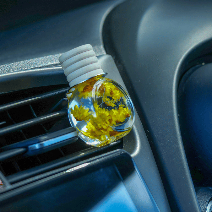 A clear car air freshener bottle with a white screw cap, filled with yellow dried flowers, resting on a car air vent.