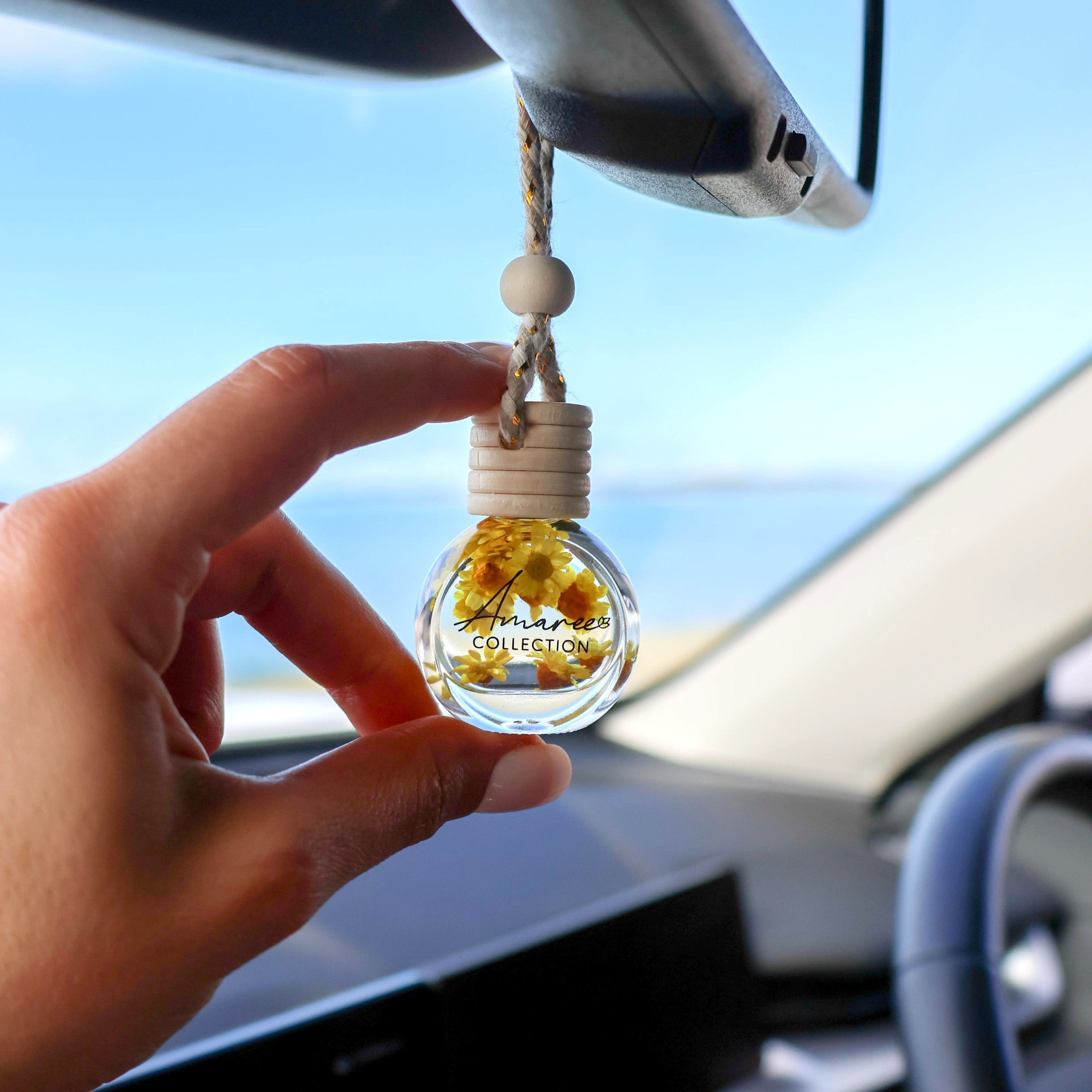 Hand holding a car air freshener with flowers inside, hanging from a car's rearview mirror.